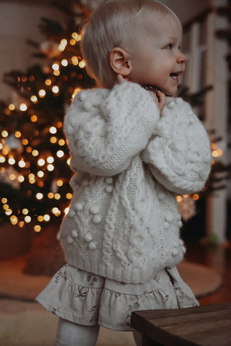 May include: A young child wearing a white knitted sweater with white pom-poms and a white and gray floral skirt. The child is standing in front of a Christmas tree with lights.