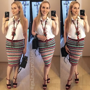 May include: A woman is wearing a white sleeveless shirt with a colorful beaded necklace, a striped pencil skirt, and red heels. She is holding a black handbag and taking a selfie.