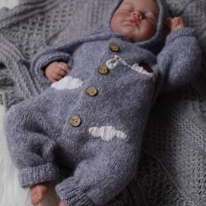Op de afbeelding: Een zachte, grijze gebreide baby romper met witte wolken borduurwerk en houten knopen. De romper heeft een capuchon en wordt gedragen door een slapende baby.