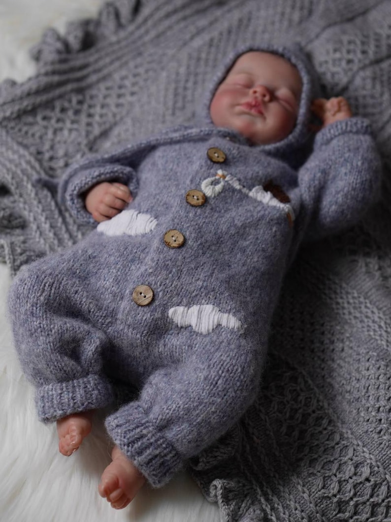 Op de afbeelding: Een zachte, grijze gebreide baby romper met witte wolken borduurwerk en houten knopen. De romper heeft een capuchon en wordt gedragen door een slapende baby.