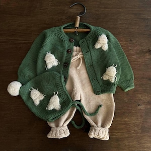 May include: A green knitted baby cardigan with white embroidered butterflies, a matching green knitted hat with a white pom-pom, and a pair of beige knitted pants.