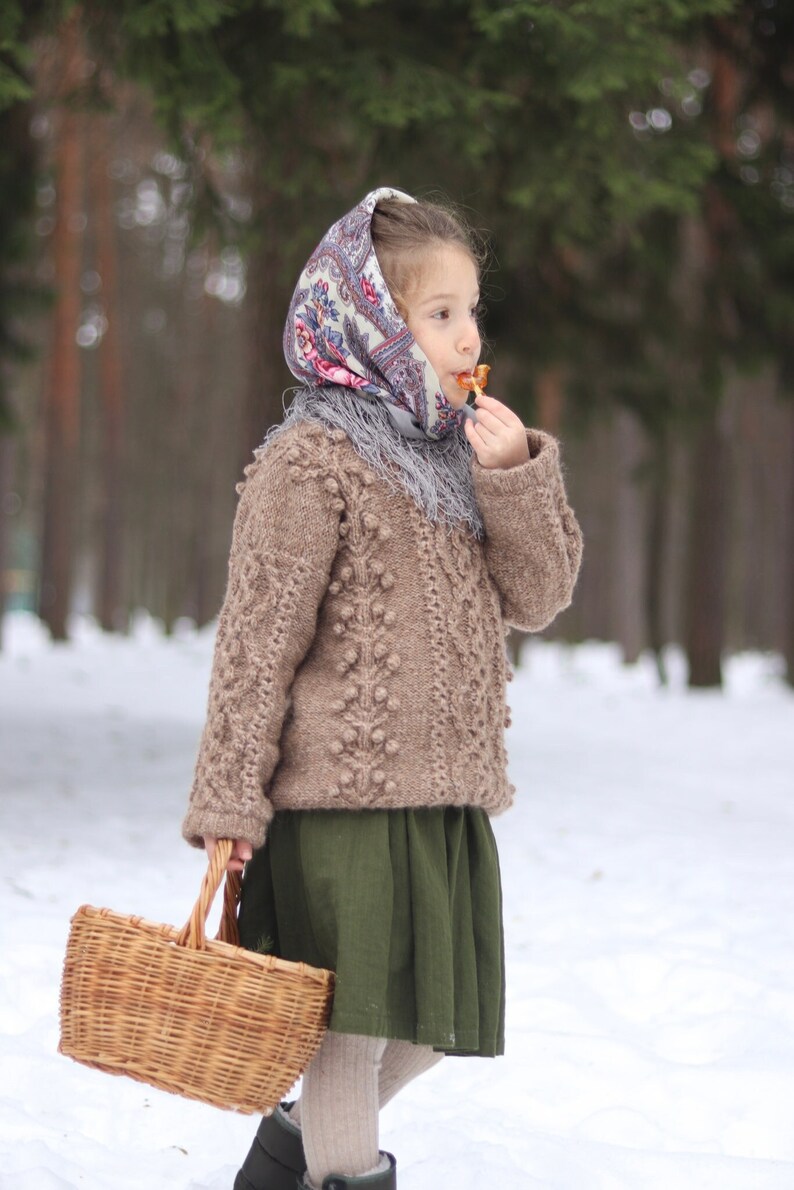 May include: A young girl wearing a brown sweater with a patterned headscarf and a green skirt holds a wicker basket in a snowy forest.