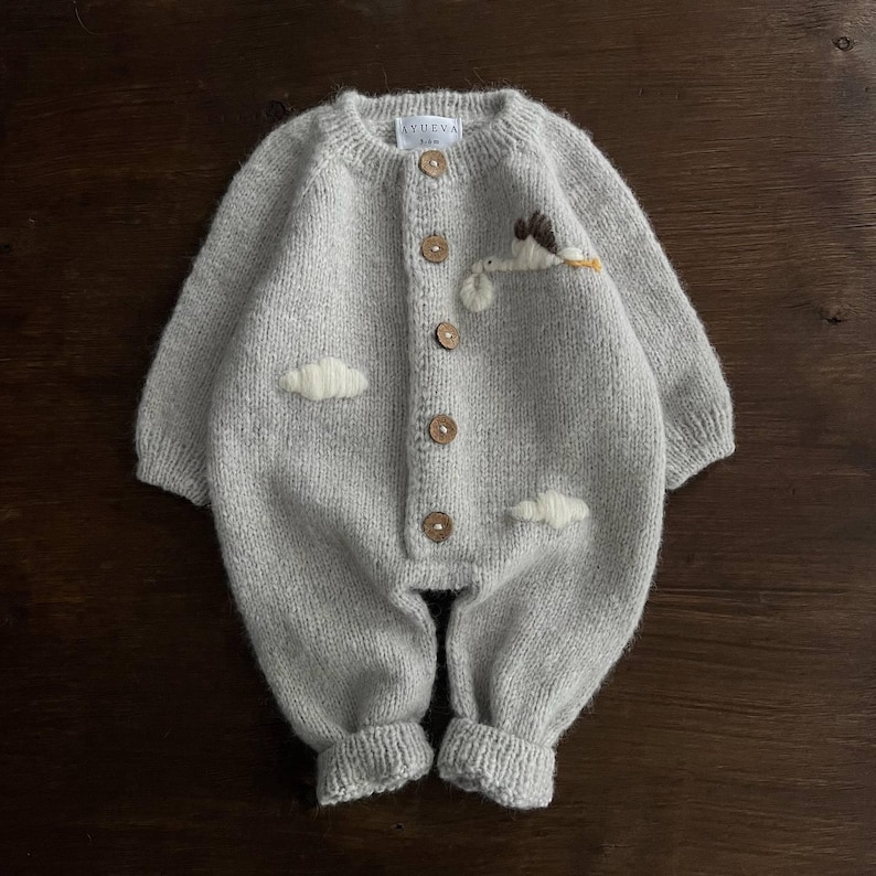 Op de afbeelding: Een grijze gebreide baby romper met een witte geborduurde ooievaar en wolken. De romper heeft een knoopsluiting aan de voorkant en is gemaakt van een zacht, comfortabel materiaal.