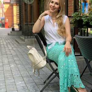 May include: A woman wearing a white sleeveless shirt, a light green crochet skirt, and gold sandals. She is sitting on a black folding chair with a white backpack hanging on the back of the chair. The woman is smiling and looking at the camera.