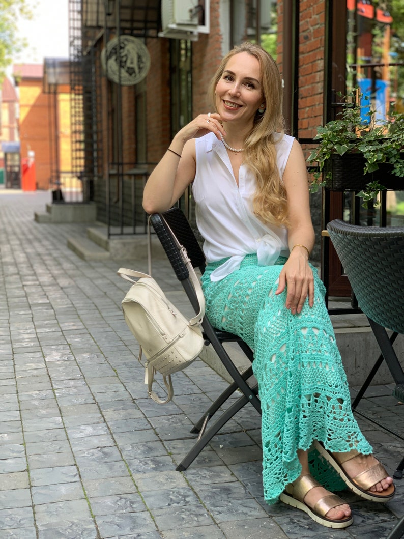 May include: A woman wearing a white sleeveless shirt, a light green crochet skirt, and gold sandals. She is sitting on a black folding chair with a white backpack hanging on the back of the chair. The woman is smiling and looking at the camera.