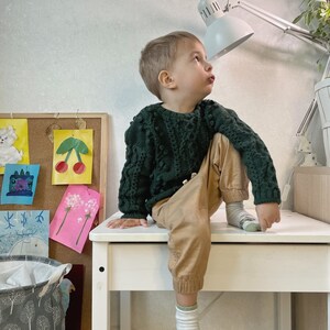 May include: A young boy wearing a dark green sweater and brown pants sits on a white table. He is looking up at a white desk lamp with a white shade. The table is in front of a corkboard with colorful artwork.