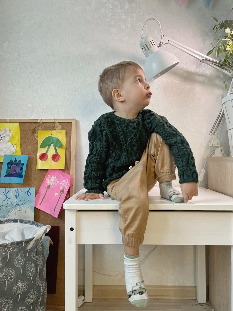 May include: A young boy wearing a dark green sweater and brown pants sits on a white table. He is looking up at a white desk lamp with a white shade. The table is in front of a corkboard with colorful artwork.