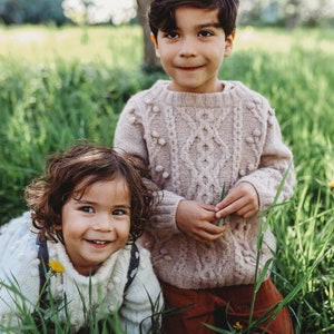 May include: Two children, a boy and a girl, are smiling at the camera. The boy is wearing a light pink cable knit sweater with white pom-poms. The girl is wearing a white cable knit sweater with a brown and white patterned strap. They are both standing in a field of green grass.