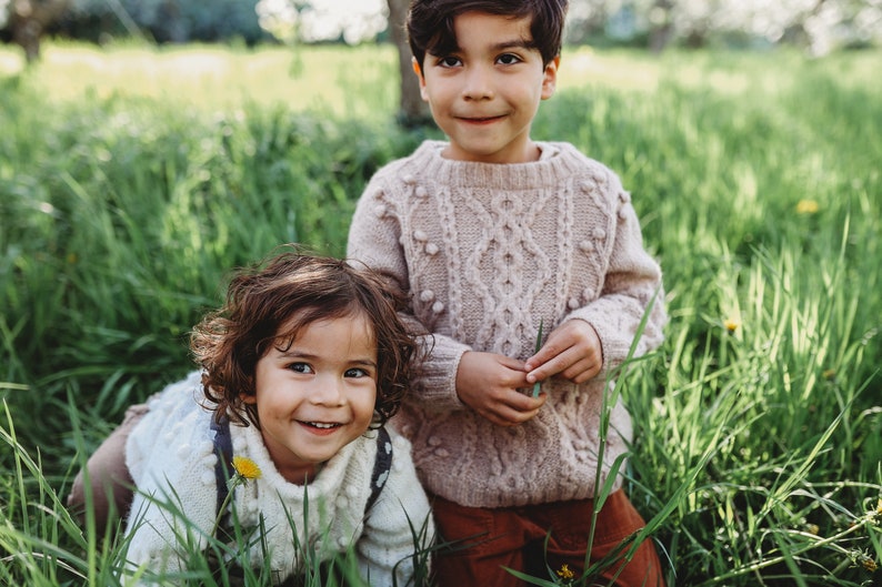 May include: Two children, a boy and a girl, are smiling at the camera. The boy is wearing a light pink cable knit sweater with white pom-poms. The girl is wearing a white cable knit sweater with a brown and white patterned strap. They are both standing in a field of green grass.