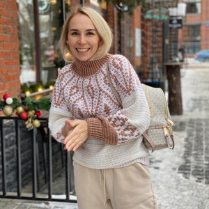 May include: A woman wearing a white and brown patterned sweater with a beige turtleneck. She is wearing beige pants and a beige backpack with a brown and white pattern. She is standing in front of a brick building with a sign that says "Coffee".