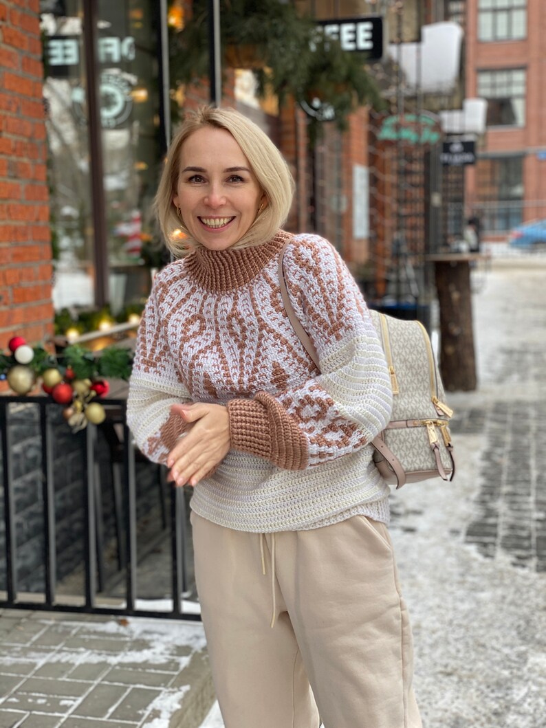 May include: A woman wearing a white and brown patterned sweater with a beige turtleneck. She is wearing beige pants and a beige backpack with a brown and white pattern. She is standing in front of a brick building with a sign that says "Coffee".