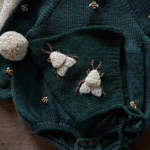 Op de afbeelding: Een donkergroene gebreide baby romper met een capuchon, met geborduurde witte motten en gele bijen. De romper heeft een pompon op de capuchon en knopen aan de onderkant.