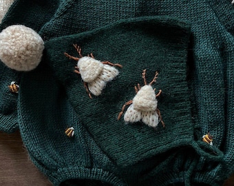 May include: A dark green knitted baby romper with a hood, featuring embroidered white moths and yellow bees. The romper has a pom-pom on the hood and buttons at the bottom.