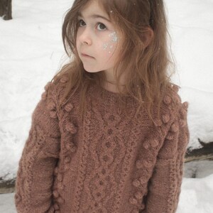 May include: A young girl wearing a brown knitted sweater with pom-poms and a reindeer headband with green leaves. She is standing in a snowy forest.