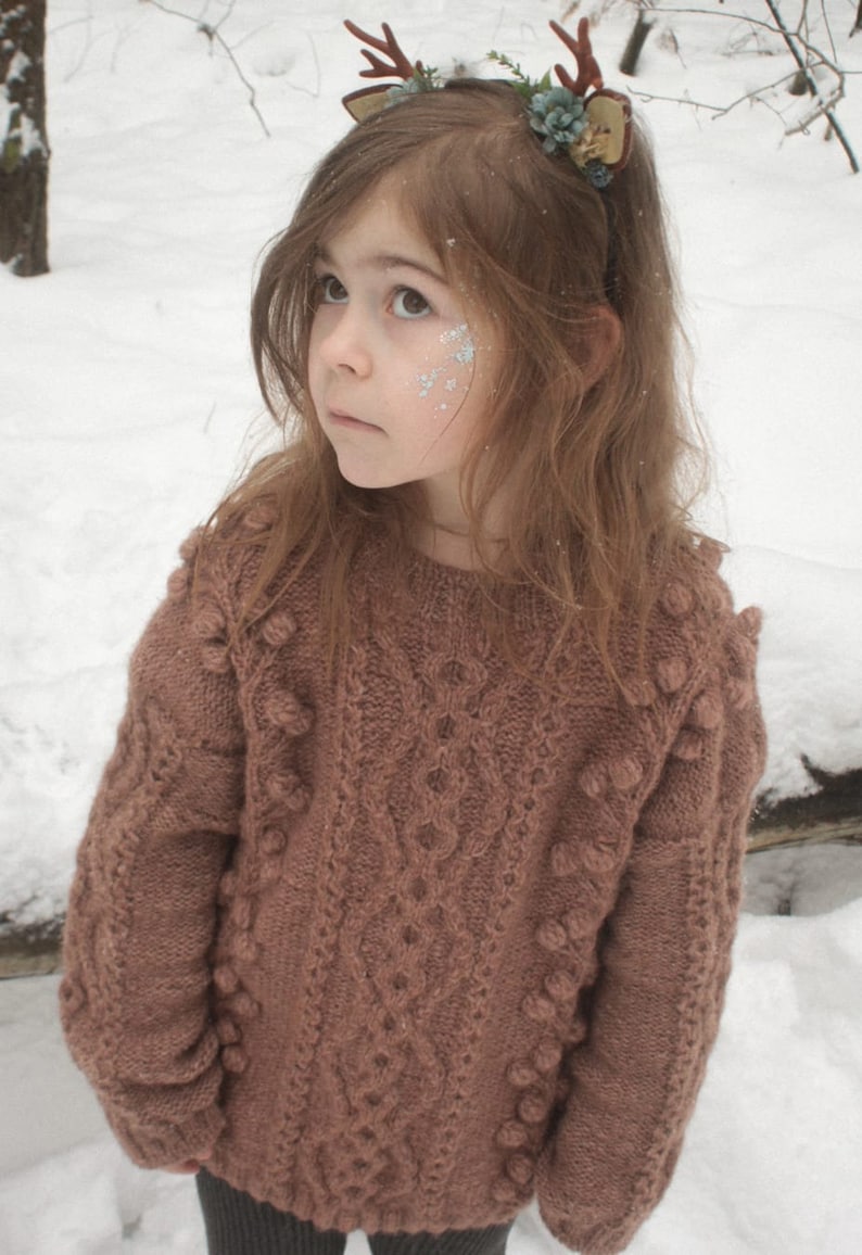 May include: A young girl wearing a brown knitted sweater with pom-poms and a reindeer headband with green leaves. She is standing in a snowy forest.