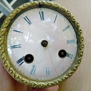 May include: Antique clock face with a light pink background and black Roman numerals. The face is cracked and has a decorative gold-colored bezel with an ornate, repeating pattern. The clock face has two small holes and a central hole.