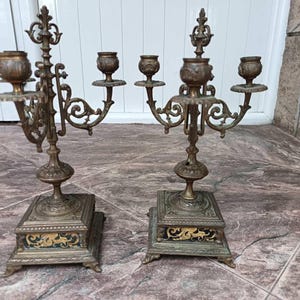 May include: A pair of ornate bronze candelabra featuring three candle holders each, supported by a central stem with intricate scrollwork. The square bases are decorated with black and gold panels, and the metal has a weathered, aged patina.