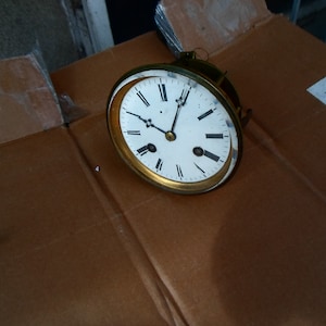 Op de afbeelding: Een vintage wijzerplaat met Romeinse cijfers en zwarte wijzers. De wijzerplaat is wit met een gouden rand en is gemonteerd op een bruin kartonnen oppervlak.