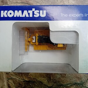May include: A yellow Komatsu bulldozer toy in a clear plastic display case. The case is inside a blue and white box with the text "Komatsu" and "The experts line".