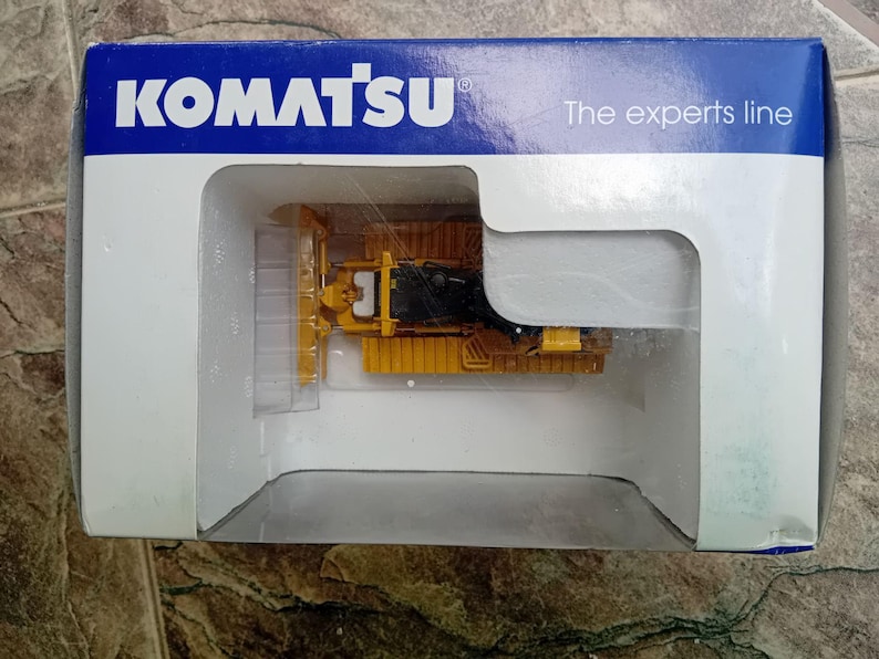 May include: A yellow Komatsu bulldozer toy in a clear plastic display case. The case is inside a blue and white box with the text "Komatsu" and "The experts line".