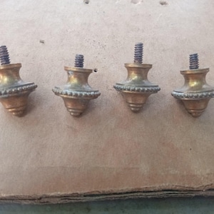 Könnte beinhalten: Vier messingfarbene Metallendstücke, jedes mit einem ausgestellten Oberteil, einem zentralen Perlenringdetail und einer spitzen Basis. Jedes Endstück enthält eine Gewindeschraube aus Metall, die oben herausragt und zur Befestigung an Möbeln dient.