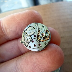 Op de afbeelding: Close-up van een zilveren uurwerk met de tekst "GIRARD-PERREGAUX" en "17 JEWELS" zichtbaar. Het uurwerk heeft een gouden tandwiel en verschillende rode juwelen.