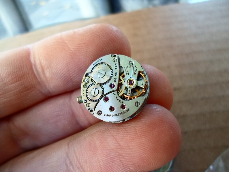 Op de afbeelding: Close-up van een zilveren uurwerk met de tekst "GIRARD-PERREGAUX" en "17 JEWELS" zichtbaar. Het uurwerk heeft een gouden tandwiel en verschillende rode juwelen.