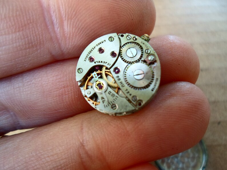 Op de afbeelding: Close-up van een zilveren uurwerk met de tekst "SIMAO-PRECISION" en "17 JEWELS" zichtbaar. Het uurwerk heeft een gouden opwindstift en verschillende tandwielen.