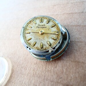 Op de afbeelding: Een close-up van een gedemonteerd horloge-wijzerplaat met een gouden wijzerplaat en zwarte wijzers. De wijzerplaat heeft de tekst "Girard Perregaux".