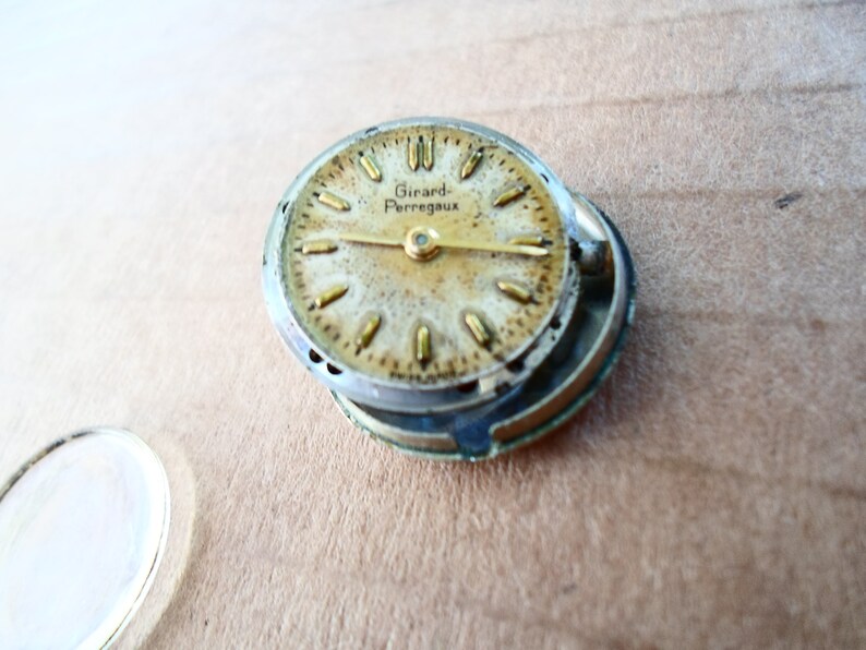 Op de afbeelding: Een close-up van een gedemonteerd horloge-wijzerplaat met een gouden wijzerplaat en zwarte wijzers. De wijzerplaat heeft de tekst "Girard Perregaux".