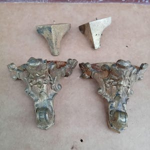 May include: Four antique gold-toned furniture feet. Two are simple triangular shapes, and two are ornate with scrollwork and floral details.