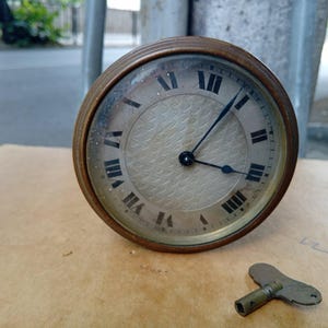 May include: An antique clock with a bronze-coloured casing and a cream-coloured face featuring black Roman numerals. The clock's hands point to the time. A small, matching key lies beside the clock. The clock's diameter is approximately 10 cm.