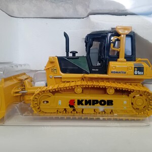 May include: A yellow toy bulldozer with a black cab and a large blade. The bulldozer has the words "Komatsu D61EX" printed on the side. The bulldozer is sitting on a white surface.