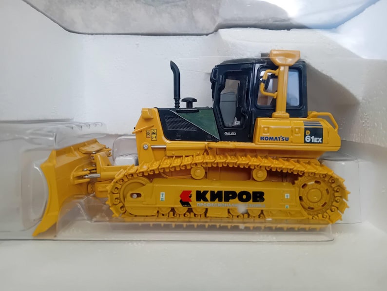May include: A yellow toy bulldozer with a black cab and a large blade. The bulldozer has the words "Komatsu D61EX" printed on the side. The bulldozer is sitting on a white surface.