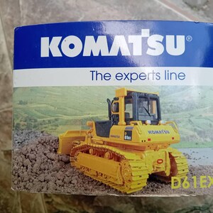 May include: A yellow Komatsu D61EX bulldozer with a black cab and a black Komatsu logo on the side. The bulldozer is on a dirt road and the box has the text "The experts line" on it.