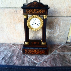 May include: A black and gold antique clock with a white face and Roman numeral markings. The clock is decorated with intricate floral patterns and sits on a marble surface.