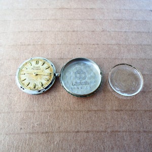 Op de afbeelding: Een zilveren wijzerplaat met de tekst "Girard Perregaux" en een zilveren horlogekast met de tekst "Girard Perregaux Automatic" en een helder glazen horlogeglas.