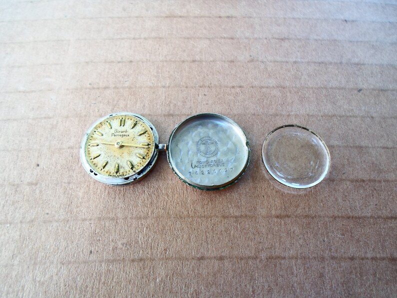 Op de afbeelding: Een zilveren wijzerplaat met de tekst "Girard Perregaux" en een zilveren horlogekast met de tekst "Girard Perregaux Automatic" en een helder glazen horlogeglas.