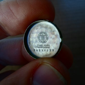 Op de afbeelding: Close-up van de achterkant van een zilveren horlogekast met de inscriptie "Fond Acier Inoxydable" en een logo met de tekst "Swiss" en een symbool van een vogel met een schild. De achterkant van de horlogekast heeft ook het nummer "70220027" gegraveerd.