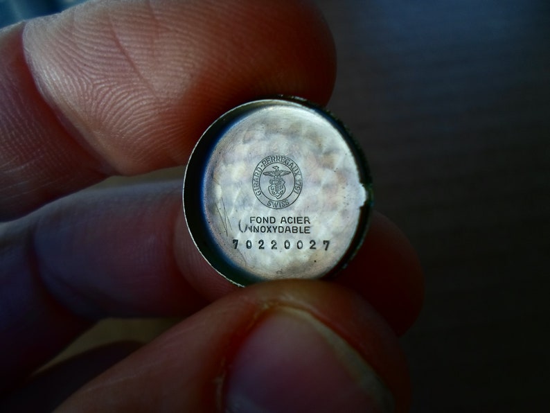 Op de afbeelding: Close-up van de achterkant van een zilveren horlogekast met de inscriptie "Fond Acier Inoxydable" en een logo met de tekst "Swiss" en een symbool van een vogel met een schild. De achterkant van de horlogekast heeft ook het nummer "70220027" gegraveerd.