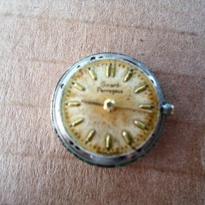 Op de afbeelding: Close-up van een rond, zilverkleurig horloge-wijzerplaat met goudkleurige wijzers en uurmarkeringen. De wijzerplaat heeft de tekst "Grand Prix" erop gedrukt.