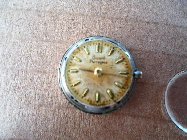 Op de afbeelding: Close-up van een rond, zilverkleurig horloge-wijzerplaat met goudkleurige wijzers en uurmarkeringen. De wijzerplaat heeft de tekst "Grand Prix" erop gedrukt.