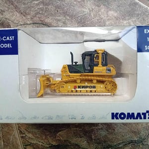 May include: A yellow die-cast model of a Komatsu D65EX-18 bulldozer with a black cab and a black blade. The model is 1:50 scale and is displayed in a clear plastic case with a blue and white background.