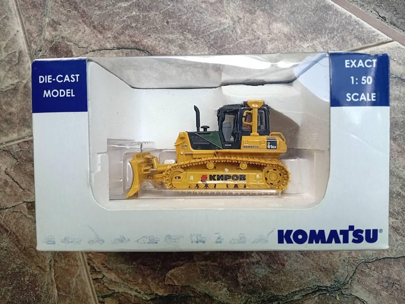 May include: A yellow die-cast model of a Komatsu D65EX-18 bulldozer with a black cab and a black blade. The model is 1:50 scale and is displayed in a clear plastic case with a blue and white background.