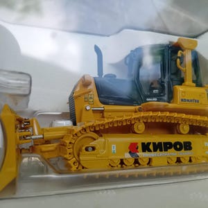 May include: A yellow toy bulldozer with a black cab and a black and yellow track. The bulldozer has the words "KOMATSU 61Ex" and "GALED" printed on the side. The bulldozer also has the words "КИРОВ ПРОФЕСИОНАЛНА ТЕХНИКА" printed on the side.
