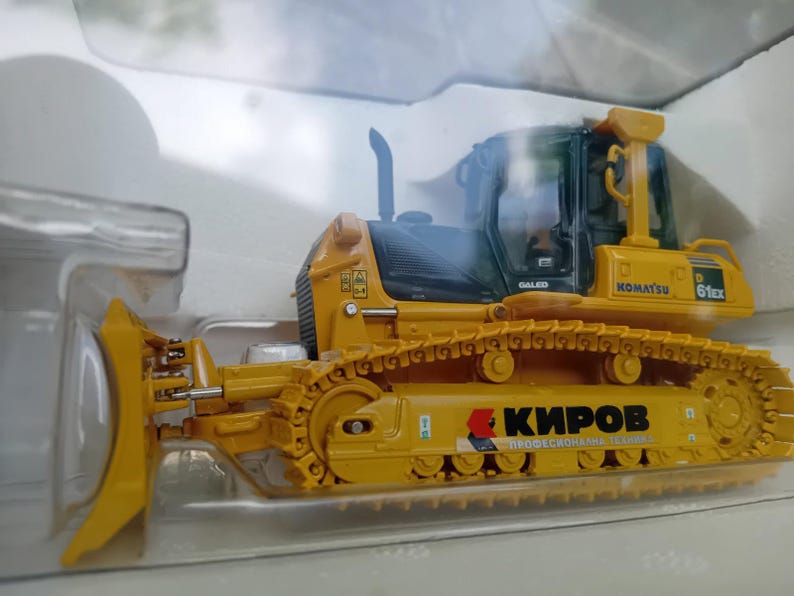 May include: A yellow toy bulldozer with a black cab and a black and yellow track. The bulldozer has the words "KOMATSU 61Ex" and "GALED" printed on the side. The bulldozer also has the words "КИРОВ ПРОФЕСИОНАЛНА ТЕХНИКА" printed on the side.