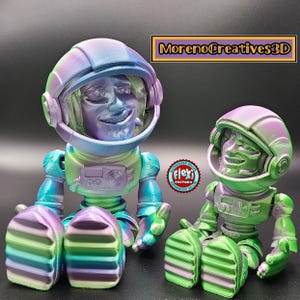 Puede incluir: Dos figuritas de astronautas iridiscentes, una más grande que la otra, con detalles verdes y morados. El astronauta más grande está sentado con las piernas cruzadas, mientras que el astronauta más pequeño está sentado con las piernas estiradas hacia adelante. Ambos astronautas tienen una expresión amistosa en sus caras.