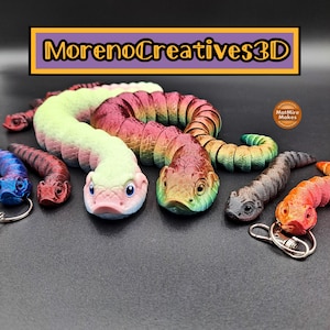 May include: A selection of vibrant, 3D-printed snake keyrings and articulated snakes. The keyrings are in blue, red, orange, and grey. The larger snakes are in green, pink, and red. The text "MorenoCreatives3D" is displayed.