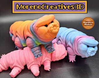 May include: Three colorful 3D printed tardigrade figurines. The first is a rainbow-colored tardigrade with an orange head. The second is a pink tardigrade. The third is a blue and pink tardigrade. The figurines are all standing on a black surface. MorenoCreatives3D and MatMire Makes are printed on the image.