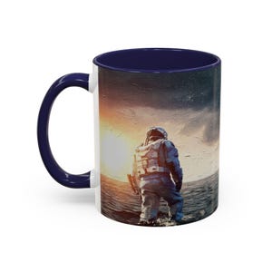 Pode incluir: Caneca de cerâmica branca com interior e alça azul marinho. A caneca apresenta um astronauta em pé na água, de frente para um pôr do sol brilhante. A obra de arte tem um estilo pictórico, com um céu estrelado ao fundo.